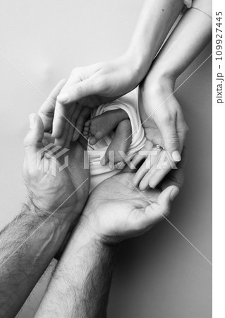 The palms of the parents, father and mother hold the legs, feet of a newborn baby. Feet, heels and toes of a newborn child close -up. Professional macro photo. Black and white.  109927445