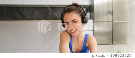 Close up portrait of brunette fitness girl, listens music in wireless headphones, smiles at camera, does workout alone at home, sits on floor and exercises 109928529