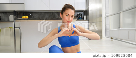 Portrait of young woman shows heart sign, sits on yoga mat, loves fitness exercises, follows online video class yoga from home, leads active lifestyle 109928684