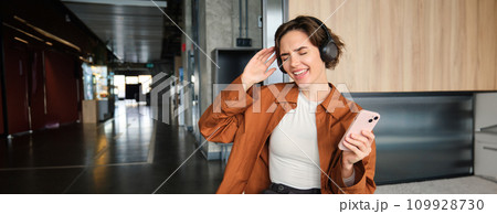 Portrait of modern young woman, office worker in lounge zone, dancing and sitting on floor of break room, enjoying her lunch, singing and listening music on smartphone 109928730