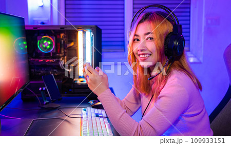 Smiling Asian woman live stream she play video game via smartphone at home neon lights living room, Gamer playing online game application on mobile phone wear gaming headphones looking to camera 109933151