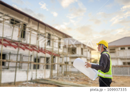 young professional engineer in protective helmet and blueprints paper at the house building construction site 109937335