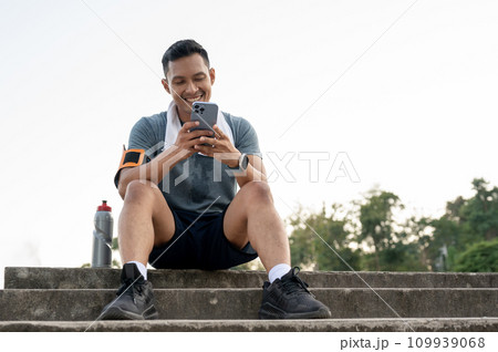 A happy man is sitting on the stairs and chatting with his friends on his phone after exercising 109939068