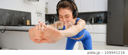Close up portrait of fit and healthy woman, smiling while doing workout exercises, listening music in wireless headphones, stretching arms, aerobics training at home, wearing blue activewear Close up portrait of fit and healthy woman, smiling while doing workout exercises, listening music in wireless headphones, stretching arms, aerobics training at home, wearing blue activewear 109941216