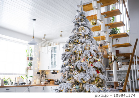 Festive Christmas decor in white kitchen, modern village interior with a snowy Christmas tree and fairy lights. New Year, Christmas mood, cozy home, Metal modular staircase. The general plan Festive Christmas decor in white kitchen, modern village interior with a snowy Christmas tree and fairy lights. New Year, Christmas mood, cozy home, Metal modular staircase. The general plan 109941374
