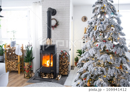 Festive white modern interior of house is decorated for Christmas and New Year in loft style with black stove, fireplace, Christmas tree. Potted plants, firewood in the woodpile Festive white modern interior of house is decorated for Christmas and New Year in loft style with black stove, fireplace, Christmas tree. Potted plants, firewood in the woodpile 109941412