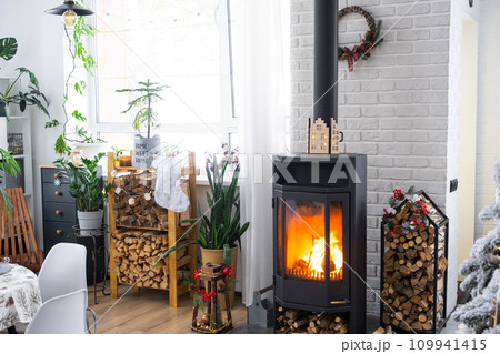 Metall black stove, burning hearth fireplace in white Festive interior of house is decorated for Christmas and New Year, Christmas tree. firewood in the woodpile, cozy and heating of home Metall black stove, burning hearth fireplace in white Festive interior of house is decorated for Christmas and New Year, Christmas tree. firewood in the woodpile, cozy and heating of home 109941415