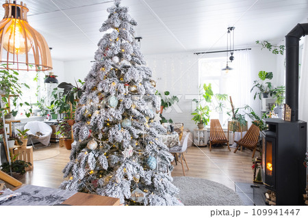 Festive white modern interior of house is decorated for Christmas and New Year in loft style with black stove, fireplace, Christmas tree. Potted plants, firewood in the woodpile 109941447