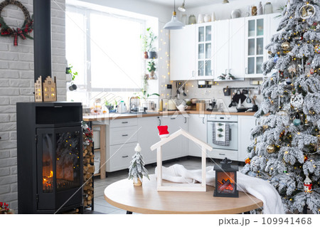Christmas decor in white kitchen with burning stove fireplace, festive mess, village interior with a snowy Christmas tree. New Year, Christmas mood, cozy home. Christmas decor in white kitchen with burning stove fireplace, festive mess, village interior with a snowy Christmas tree. New Year, Christmas mood, cozy home. 109941468