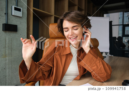 Portrait of young office employee, woman working in conference room, dancing while sitting with wireless headphones, listening to music Portrait of young office employee, woman working in conference room, dancing while sitting with wireless headphones, listening to music 109941758