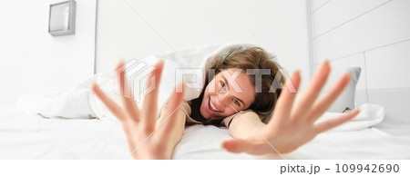 Beautiful woman lying in bed, stretching her arms towards camera and laughing, waking up in morning in happy mood Beautiful woman lying in bed, stretching her arms towards camera and laughing, waking up in morning in happy mood 109942690
