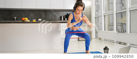 Portrait of active and healthy young woman, doing squats with resistance band, workout at home, listening music in wireless headphones Portrait of active and healthy young woman, doing squats with resistance band, workout at home, listening music in wireless headphones 109942850
