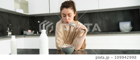 Portrait of young beautiful woman in bathrobe, eating cereals for breakfast, leans on kitchen worktop, looking at her morning meal 109942998