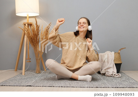 Overjoyed cheerful woman with brown hair wearing beige casual clothing and headphones hearing favorite song on radio dancing being in good mood sitting on floor in light room Overjoyed cheerful woman with brown hair wearing beige casual clothing and headphones hearing favorite song on radio dancing being in good mood sitting on floor in light room 109948093