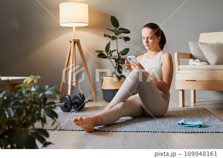 Sport and fitness online. Yoga with smartphone. Athletic workout at home. Fitness app for training. Sporty woman wearing beige tracksuit sitting on floor using mobile cell phone Sport and fitness online. Yoga with smartphone. Athletic workout at home. Fitness app for training. Sporty woman wearing beige tracksuit sitting on floor using mobile cell phone 109948161