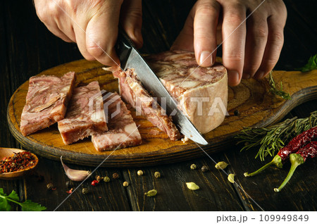 The cook cuts the brawn with a knife on the kitchen board before preparing sandwiches. Idea for a delicious snack with headcheese and aromatic spices. 109949849