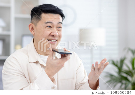 Close-up photo of a young smiling Asian man sitting at home talking on the phone through the speakerphone, recording the conversation, asking in the application. Close-up photo of a young smiling Asian man sitting at home talking on the phone through the speakerphone, recording the conversation, asking in the application. 109950584