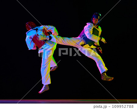 Dynamic image of young men, taekwondo athletes in kimono and helmets training against black background in neon light. Martial arts, combat sport Dynamic image of young men, taekwondo athletes in kimono and helmets training against black background in neon light. Martial arts, combat sport 109952788