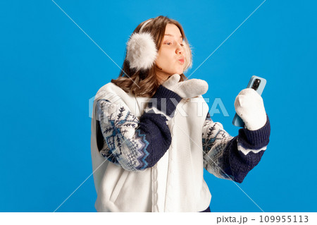 Portrait of young girl, student blows kiss to her phone camera while communicating via video conference against blue studio background. Portrait of young girl, student blows kiss to her phone camera while communicating via video conference against blue studio background. 109955113