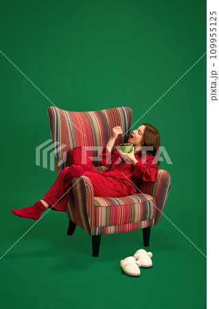 Full length portrait of beautiful young girl lying in red silk pajamas on retro armchair and eats delicious sweet cereal against green studio background. 109955125
