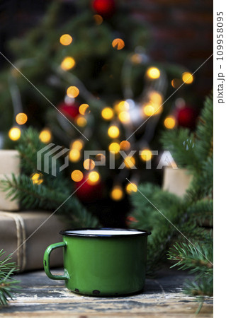 Coffee in a tin mug on the rustic table with wrapped gifts, Christmas tree lights and spruce branches in the background 109958095