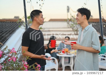 Two young handsome men having drinks and chatting at the rooftop party in the evening. Two young handsome men having drinks and chatting at the rooftop party in the evening. 109967217