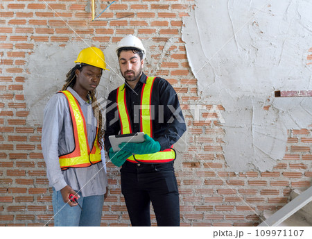 Two construction workers checking work schedule on tablet computer. Wear hardhat and safety vest, stand in front of a brick wall in construction site. 109971107