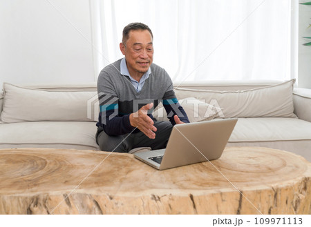 Old man relaxing comfortably on a sofa, immersed in operating a laptop. Evident focus and concentration in a cozy domestic backdrop. 109971113