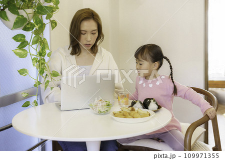 キッチンでパソコン作業をする茶髪の若いママとテーブルでおにぎりを食べる女の子 キッチンでパソコン作業をする茶髪の若いママとテーブルでおにぎりを食べる女の子 109971355