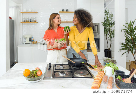 Two women are in the kitchen, happily working together to prepare a meal, chopping vegetables and mixing ingredients. Two women are in the kitchen, happily working together to prepare a meal, chopping vegetables and mixing ingredients. 109971774