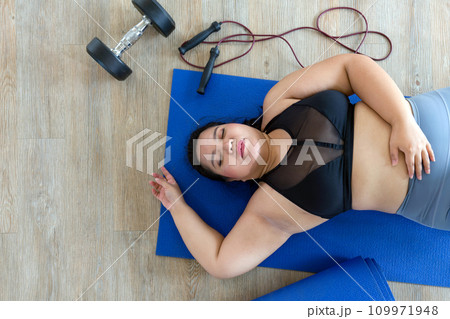 A plus-size woman lies exhausted on a blue mat, taking a breather after a vigorous workout session with dumbbell and jump rope. Top view A plus-size woman lies exhausted on a blue mat, taking a breather after a vigorous workout session with dumbbell and jump rope. Top view 109971948