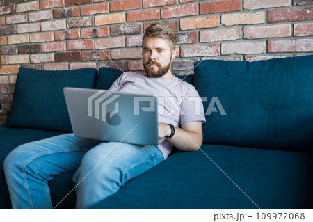 Millennial man sitting on sofa at home working on laptop online and using internet. Smiling and happy mood, freelancer and free leisure time, relaxed and modern job lifestyle concept. Copy space and 109972068