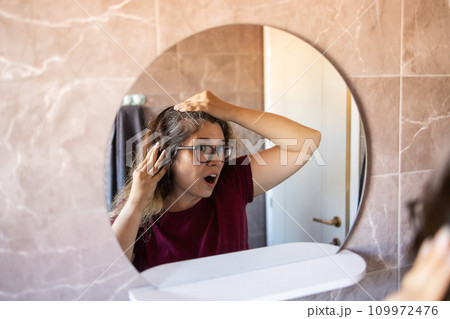 Gray haired surprised caucasian middle aged woman looking at grey hair head in mirror reflection. Early gray hair concept 109972476