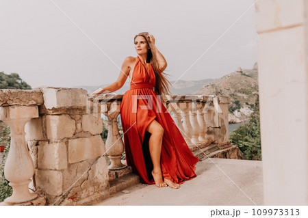 Woman travel portrait. Happy woman with long hair looking at camera and smiling. Close up portrait cute woman in a res long dress posing on backdrop of old travel city near sea 109973313