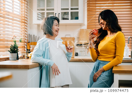 A smiling mother and her adult daughter, an attractive young woman, stand together in the kitchen, holding an apple. Their bond radiates happiness and family togetherness. 109974475