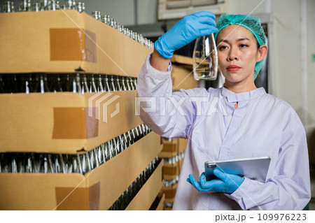 A QC woman working in a beverage factory utilizes a digital tablet to check products on the conveyor belt. She ensures quality control while examining the bottling line for liquid manufacturing. A QC woman working in a beverage factory utilizes a digital tablet to check products on the conveyor belt. She ensures quality control while examining the bottling line for liquid manufacturing. 109976223