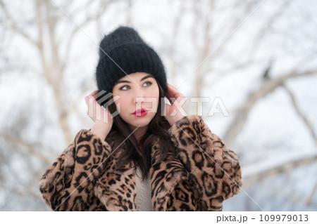 Winter portrait of young woman with long hair and red plump lips thoughtful looking away. Romantic brunette female dressed in fur coat with leopard pattern raised hands and straightens knitted hat 109979013