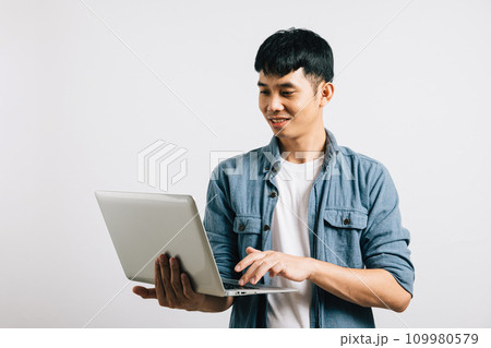 Portrait of confident Asian businessman, smiling and typing on a laptop keyboard, engaged in a successful online email or chat isolated on white background, showcasing his enthusiasm for technology. 109980579