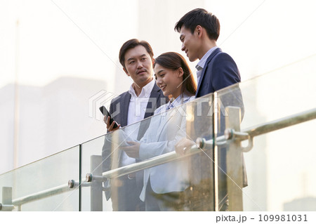 asian business people looking at mobile phone chatting outdoors 109981031