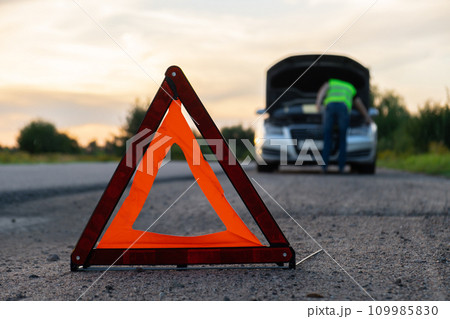 Broken silver luxury car emergency accident. Man driver installing red triangle stop sign on road. Sport automobile turned on blinkers technical problems on the road. Safety procedure vehicle broken Broken silver luxury car emergency accident. Man driver installing red triangle stop sign on road. Sport automobile turned on blinkers technical problems on the road. Safety procedure vehicle broken 109985830