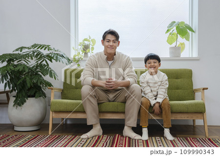 ソファに並んで座るカメラ目線で笑顔の親子　ポートレート　30代の父親と小学1年生の息子 109990343