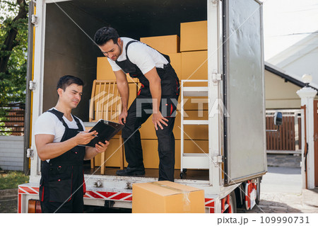 Mover workers in uniform unload boxes checking checklist with a clipboard at the truck. Professional delivery service ensures smooth relocation and efficient shipping. Moving day concept Mover workers in uniform unload boxes checking checklist with a clipboard at the truck. Professional delivery service ensures smooth relocation and efficient shipping. Moving day concept 109990731