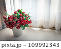 Roses in a vase on a table next to a window lined with white curtains 109992343
