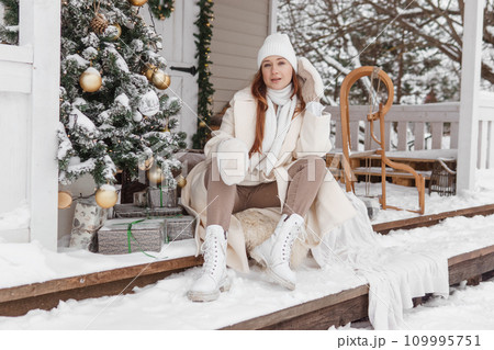 A woman in white winter clothes on the veranda of a country house decorated for the Christmas holidays. Wooden house. A woman in white winter clothes on the veranda of a country house decorated for the Christmas holidays. Wooden house. 109995751