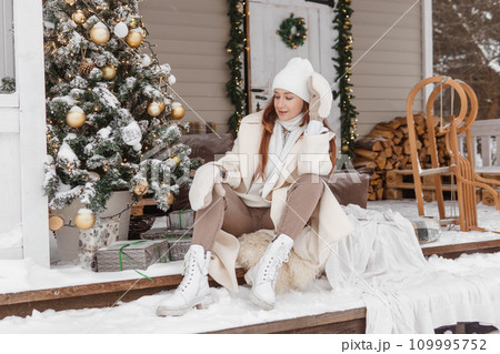 A woman in white winter clothes on the veranda of a country house decorated for the Christmas holidays. Wooden house. A woman in white winter clothes on the veranda of a country house decorated for the Christmas holidays. Wooden house. 109995752