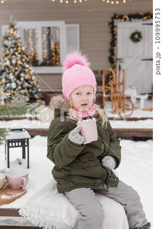 A little girl in winter clothes with a cup of hot cocoa with marshmallows, in the courtyard of a country house decorated for the Christmas holidays. Christmas holidays. A little girl in winter clothes with a cup of hot cocoa with marshmallows, in the courtyard of a country house decorated for the Christmas holidays. Christmas holidays. 109995753
