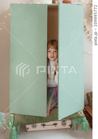 A little red-haired girl is sitting in the closet of a children's room decorated for the Christmas holidays. The girl is holding a gift box in her hands A little red-haired girl is sitting in the closet of a children's room decorated for the Christmas holidays. The girl is holding a gift box in her hands 109995772