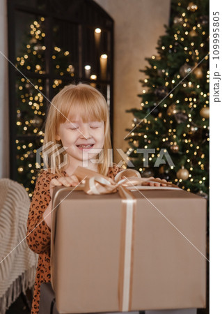 A little blonde girl in a festive interior decorated in a New Year's style. A child holds a Christmas gift in a craft package. A little blonde girl in a festive interior decorated in a New Year's style. A child holds a Christmas gift in a craft package. 109995805