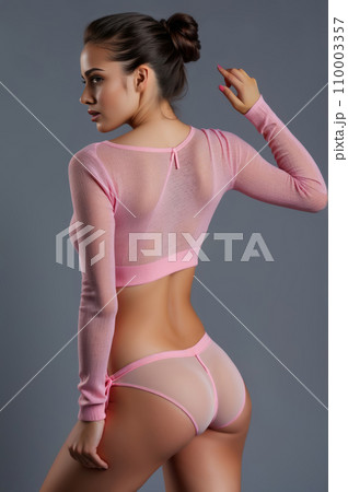 Provocative young woman posing in pink sweater and sexy pink lingerie on studio gray background. Slender figure and abdomen partially revealed. 110003357