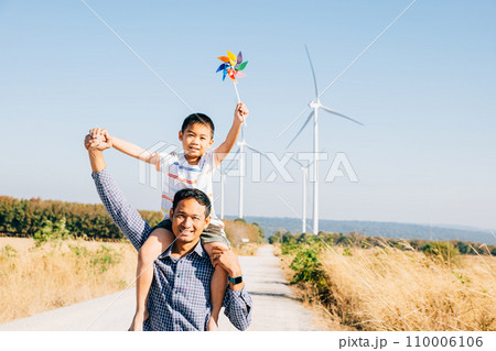 Father holds daughter with pinwheel enjoying. Family bonding near windmills signifies global innovation in renewable energy. A joyful moment in the community's pursuit of happiness. Father Day 110006106